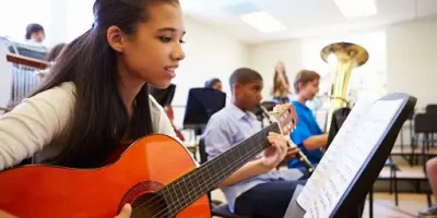 Female Pupil Playing Guitar In High School Orchestra Reading Music Sheet